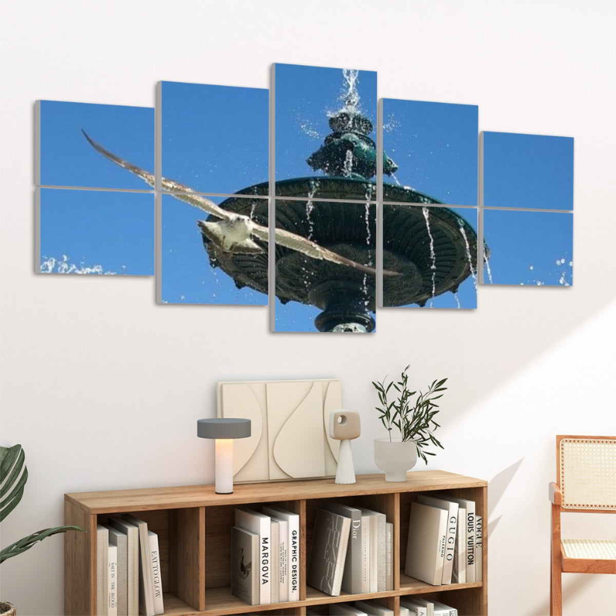 Ten panels mounted above a wooden console in a living room. The vivid blue sky, detailed bronze fountain spout, and white seagull flying overhead stand out against a neutral wall, with soft natural light illuminating fragments of the image